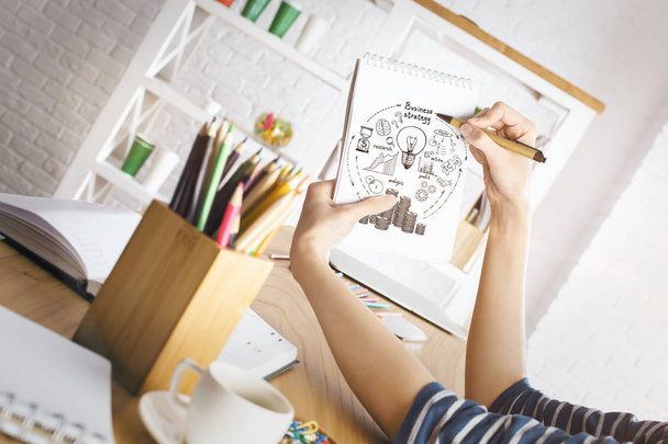 Woman drawing business sketch in spiral notepad placed on wooden office desk with coffee cup, supplies and other items. Idea concept - Foto, Imagen
