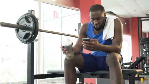 Sedí v tělocvičně, sportovec pít protein koktejl a posouvání na telefonu - Záběry, video