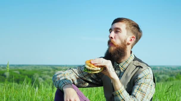 A young bearded man eating a hamburger outdoors. Sits on a meadow in a picturesque place In sring sunny day - Záběry, video