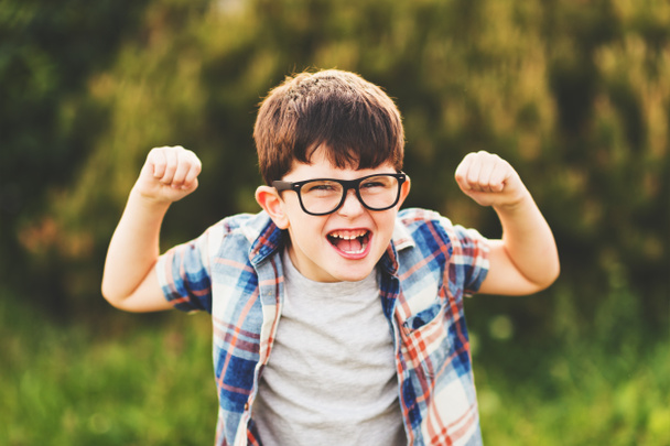 Erős és okos kisfiú vicces arc kifejezése, játék a szabadban, szemüveg és kék kockás inget visel - Fotó, kép