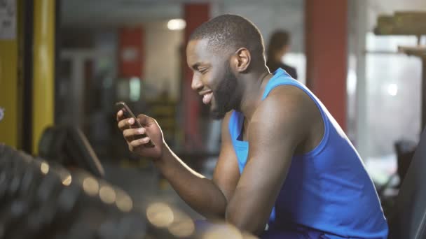 Young man typing message on smartphone with smile on face, break during workout - Video, Çekim