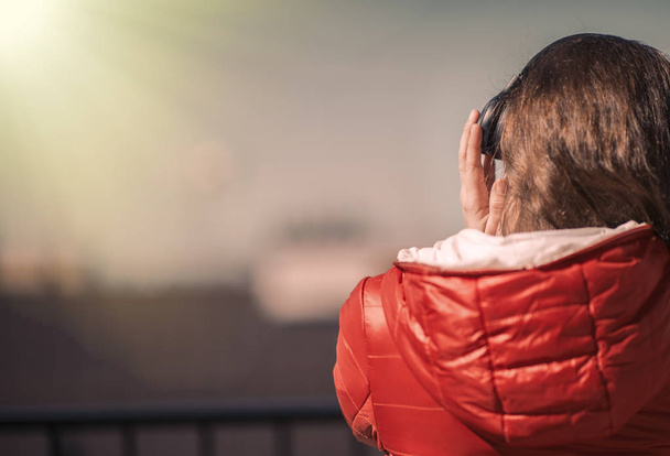 Beautiful girl in a red jacket and headphones. A girl on the balcony listening to music and is singing - Fotó, kép