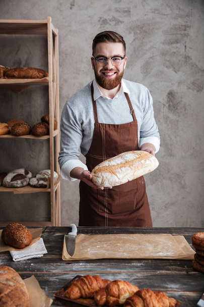 Cheerful young man baker standing at bakery holding bread - Foto, Imagem