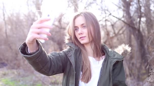 ブロンドの髪と美しいヨーロッパの女の子はジャケットに身を包んだ、公園で、ソーシャル メディアのウェブサイトにアップロードした後、selfie を取る。屋外。Selfie. - 映像、動画