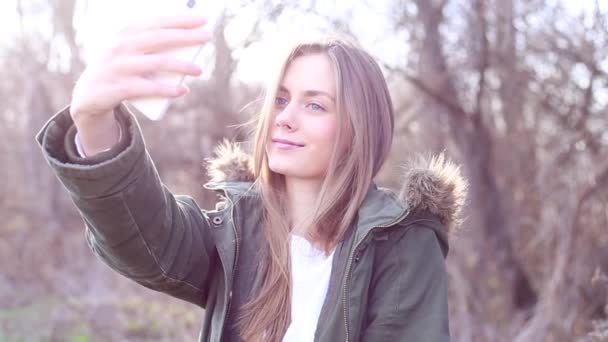 ブロンドの髪を持つ美しい少女のジャケットの服を着て、公園で、ソーシャル メディアのウェブサイトにアップロードした後、selfie を取る - 映像、動画