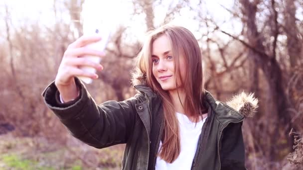 Menina europeia bonita com cabelo loiro vestido com uma jaqueta e tirar uma selfie no parque e depois de carregado para um site de mídia social
 - Filmagem, Vídeo