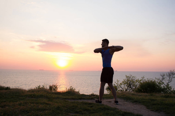 Young male stretching before fitness training session at the park. Healthy young man warming up outdoors. Sunset or sunrise in the sea or ocen on background - Valokuva, kuva