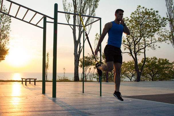 workout with suspension straps In the outdoor gym, strong man training early in morning on the park, sunrise or sunset in the sea background  - Foto, imagen