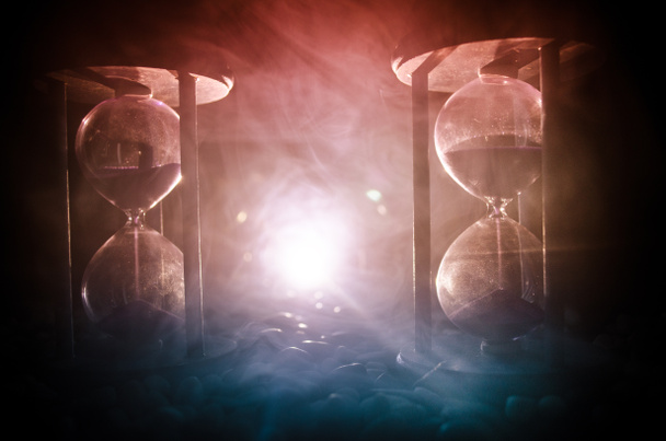 Concept du temps. Sable passant à travers les ampoules en verre d'un sablier mesurant le temps qui passe car il compte jusqu'à une date limite. Silhouette de sabliers en fumée sur fond sombre. Avec des fleurs
 - Photo, image