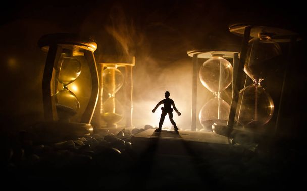 Tijd concept. Silhouet van een man die tussen hourglasses met rook en licht op een donkere achtergrond. - Foto, afbeelding