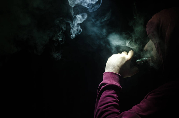 Vaping man holding a mod. A cloud of vapor. Black background. Vaping an electronic cigarette with a lot of smoke - Foto, imagen