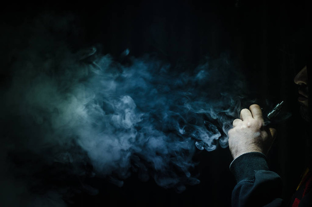 Vaping man holding a mod. A cloud of vapor. Black background. Vaping an electronic cigarette with a lot of smoke - Valokuva, kuva