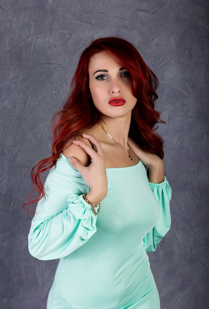 Portrait of beautiful redhead young woman with big boobs in a turquoise dress on a gray background - Фото, зображення