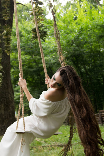 meisje met lang haar in witte jurk is rope swing in fores rijden - Foto, afbeelding