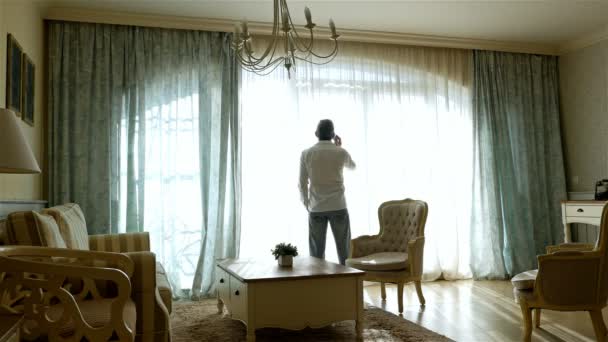 Hombre preocupado yendo y viniendo por la ventana de un acogedor apartamento, hablando por teléfono celular
 - Metraje, vídeo
