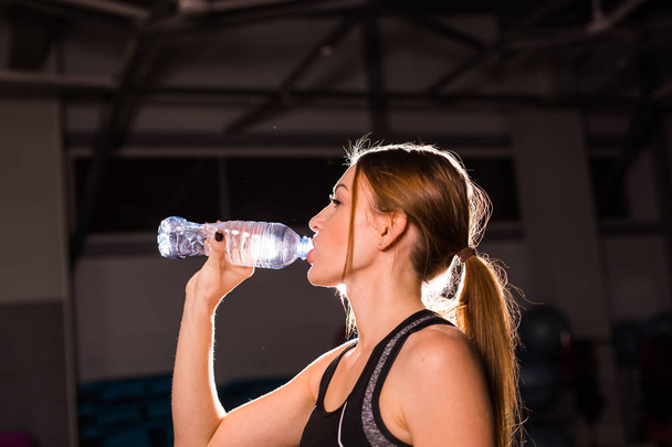 Mujer fitness bebiendo agua de la botella. Musculosa joven en el gimnasio tomando un descanso del entrenamiento
. - Foto, Imagen