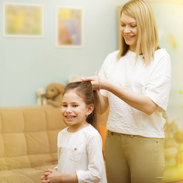 Mãe está trançando o cabelo de sua filha jovem de pé na sala de crianças
.  - Foto, Imagem