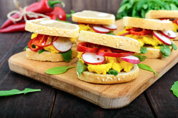 Sandwich met roerei, rucola, radijs, paprika kapi en lijnzaad op een snijplank op een donkere houten achtergrond. Snel en lekker ontbijt. - Foto, afbeelding