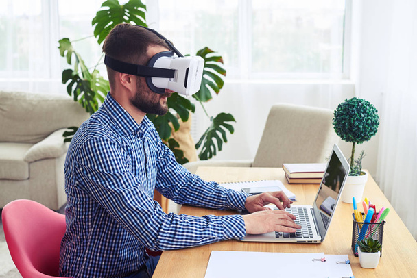 Man in VR glasses working with laptop sitting at table - Photo, Image