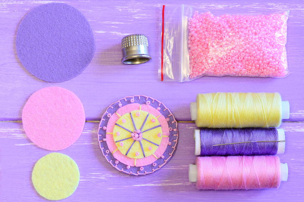Felt flower, colored felt circles, thread spools, needle, thimble, pink beads on a wooden table. Sewing felt flower tutorial. Simple round felt flower crafts for kids. Top view  - Photo, Image