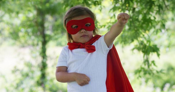 in a beautiful and happy sunny day , a little girl dressed as a super hero makes expressions and try to fly in the colorful nature , the child laughs happy . concept of happiness , love and nature. - Photo, Image
