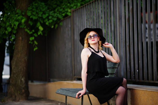 Blonde woman on black dress, sunglasses, necklaces and hat sitti - Fotó, kép