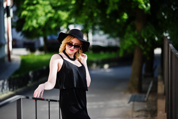 Blonde woman on black dress, sunglasses, necklaces and hat posed - Fotó, kép