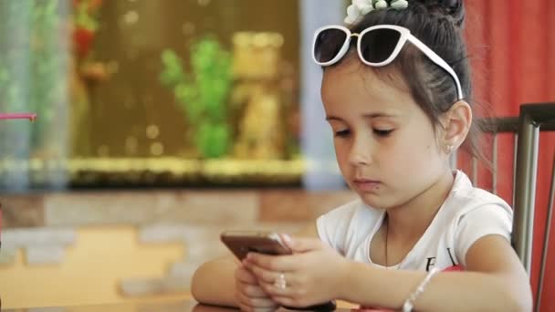 Malá dívka dítě bruneta latina posezení v kavárně, na smartphone ve hře, rozhořčený, nešťastný - Záběry, video