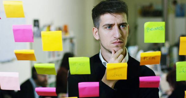 Portrait of a smiling man in a thoughtful and creative agency with colorful notes and planning hanging behind the glass and the working people. Concept: team work, agency, creative, business - Photo, image