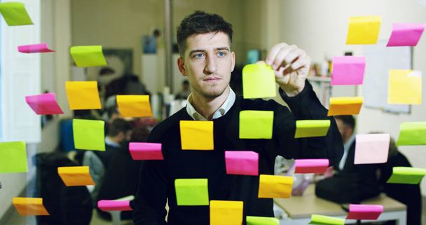 Portrait of a smiling man in a thoughtful and creative agency with colorful notes and planning hanging behind the glass and the working people. Concept: team work, agency, creative, business - Foto, Imagem