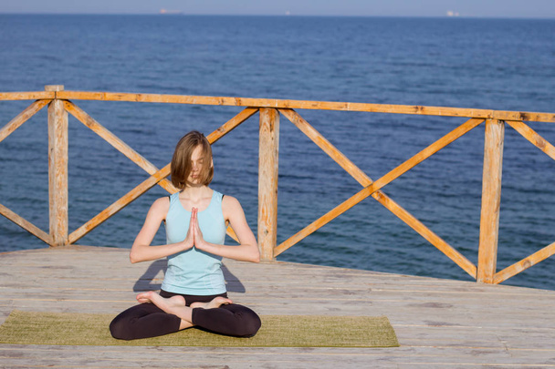 Young sexy woman doing yoga exercises on the wooden pier against sea background and blue sky - Photo, image