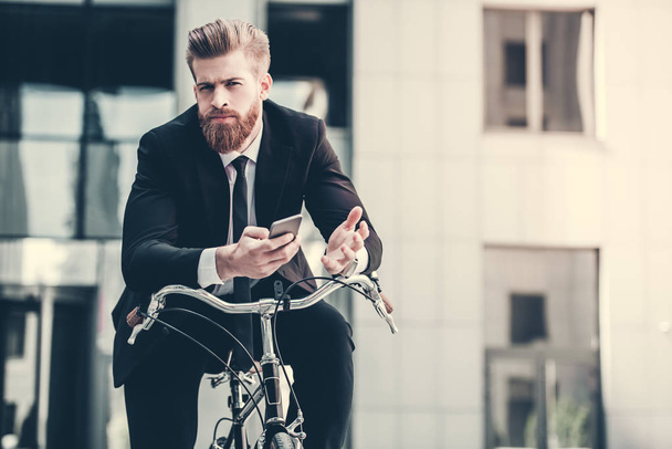 Handsome bearded businessman in classic suit is using a smart phone and looking at camera while riding bicycle in city - Zdjęcie, obraz