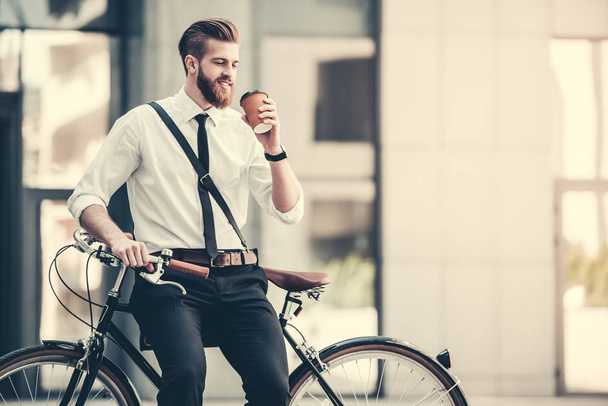 Handsome bearded businessman in classic suit and with bag is drinking coffee and smiling while riding bicycle in city - 写真・画像