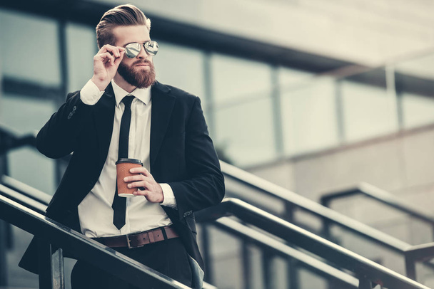 Handsome bearded businessman in classic suit and sun glasses is holding a cup of coffee while standing on the stairs in the city center - Foto, Imagen