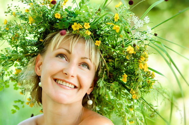 Joven chica feliz en corona de hierbas y flores. Día de verano en un prado
. - Foto, Imagen