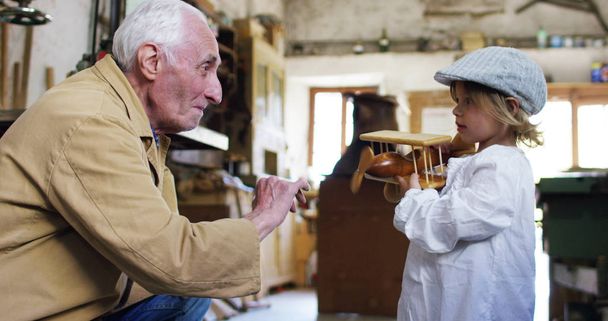in an old carpentry shop a child and grandfather playing with a wooden airplane concept of tradition that continues over time and surprise - Fotoğraf, Görsel