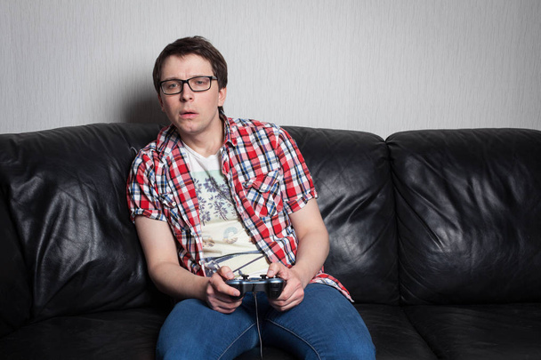 Young guy with glasses and red shirt playing video games on the joystick, sitting on a black leather sofa - Fotó, kép