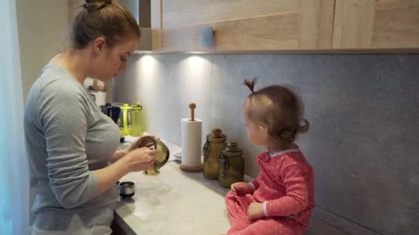 Happy young mother cooks with a child who sits on a table at home kitchen - Felvétel, videó