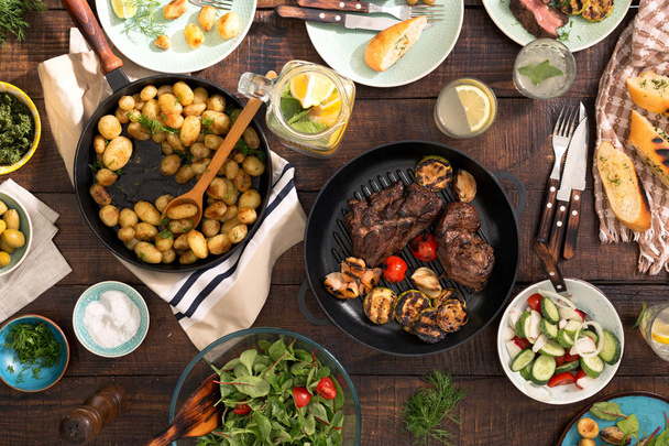 Dinner table with grilled steak, vegetables, potatoes, salad, sn - Foto, afbeelding