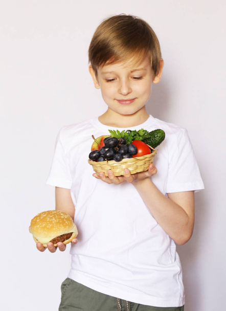 ein blonder Junge wählt zwischen einem Burger und frischem Gemüse und Obst. - Foto, Bild