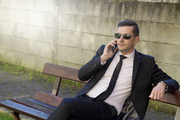 Young man in suit sitting in park talking with mobile phone - 写真・画像