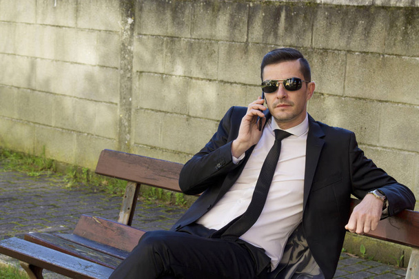 Young man in suit sitting in park talking with mobile phone - Photo, image