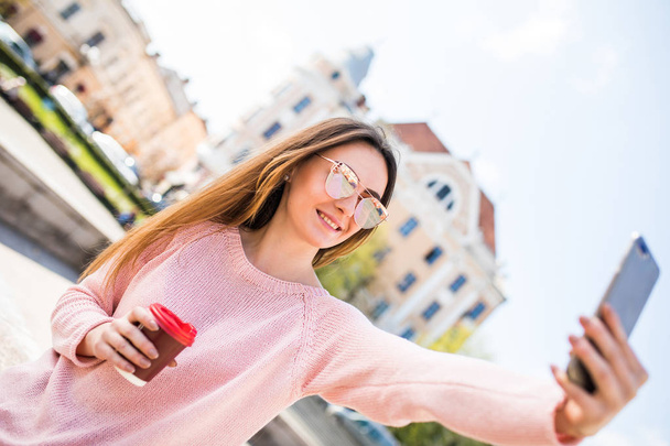 Cheerful woman with amazing smile wearing modern outfit makes selfie with coffee while walk on old town square - Φωτογραφία, εικόνα