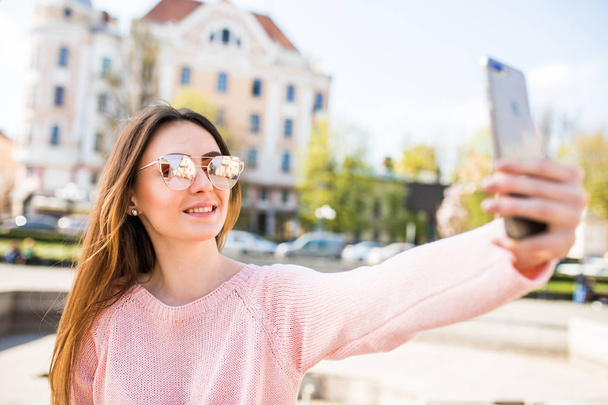 Молодая девушка делает селфи из рук с телефоном на летней улице города. Концепция городской жизни
. - Фото, изображение