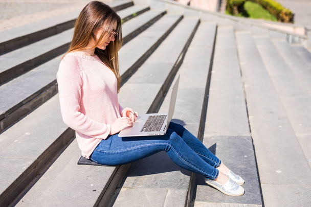 Porträt einer glücklichen jungen Frau, die auf der Treppe der Stadt sitzt und Laptop benutzt - Foto, Bild