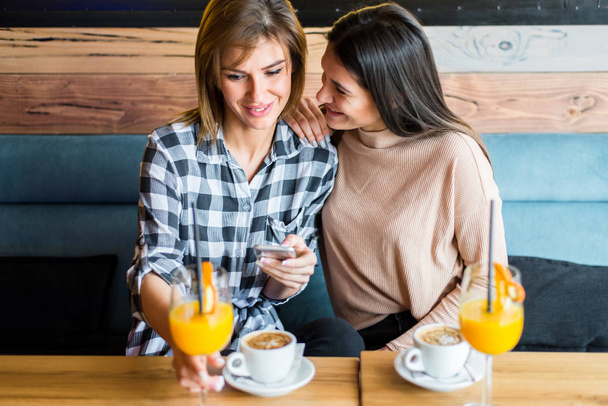 Kaksi nuorta naisystävää kahvilassa, juomassa kahvia ja mehua, katsomassa jotain matkapuhelimesta.
 - Valokuva, kuva
