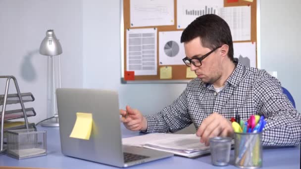 Der geschäftige Mann füllt am Arbeitsplatz Papierformulare aus und blickt auf den Laptop-Monitor - Filmmaterial, Video
