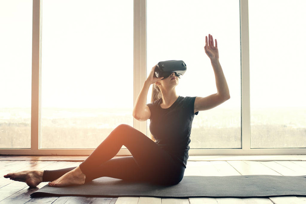 Una joven hermosa mujer en gafas de realidad virtual hace aeróbicos remotamente. Concepto de tecnología futura. Tecnología de imagen moderna. Clases en deportes individuales remotamente
. - Foto, imagen