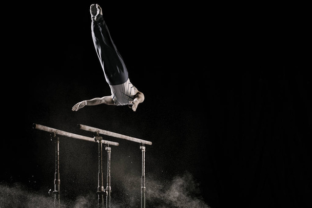 Male athlete performing difficult exercise on gymnastic parallel bars with talcum powder. Isolated on black. - Photo, Image