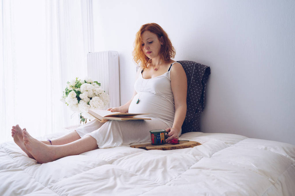 Young pregnant woman relaxing on the bed with a book - Фото, изображение